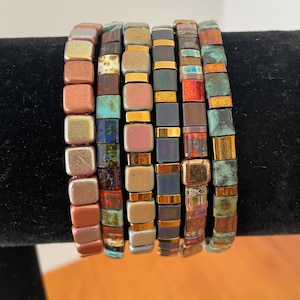 May include: Six beaded bracelets with square, iridescent tiles in shades of copper, gold, teal, and navy blue. The bracelets are displayed on a black surface. Each bracelet has a different color scheme.