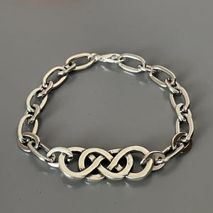 May include: A silver bracelet featuring an infinity symbol design. The bracelet is composed of oval-shaped links connected by a clasp. The central design element is three intertwined infinity symbols.