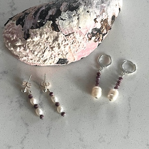 May include: An overhead shot of two pairs of earrings and a large seashell. One pair of earrings has a pearl and purple bead design with a post back. The other pair has a pearl and purple bead design with a hoop back. The seashell is white, pink, and black.