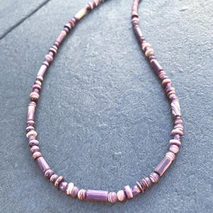 May include: A handmade necklace featuring a mix of purple and white beads. The necklace has a variety of bead shapes, including small round beads and larger cylindrical beads. The beads are strung together on a thin, dark-colored cord.