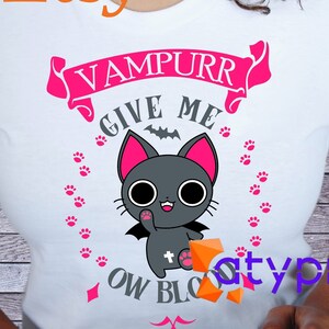 Puede incluir: Camiseta blanca con un gráfico rosa y gris de un gato vampiro con el texto "Vampurr Give Me Ow Blood".