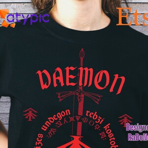 May include: A black t-shirt with a red graphic design featuring a sword and the word "DAEMON" in a stylized font. The design also includes the text "izes undegon rebzi kostol" in a smaller font. The text "Designed by RaDoNeLaJ" is also included in the graphic.