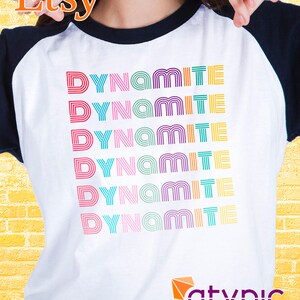 May include: A white and navy blue baseball tee shirt with the word "Dynamite" repeated six times in rainbow colors. The word is in a retro font style.
