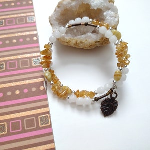 May include: A double-strand bracelet with white and brown beads and a small leaf charm. The bracelet is on a white surface with a rock behind it.