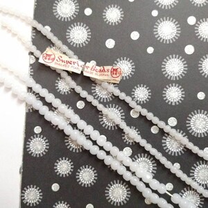 May include: Three strands of white pearl beads on a black and white patterned background. The beads are labeled "Superior Beads" and "Simulated Pearl Made in Japan."