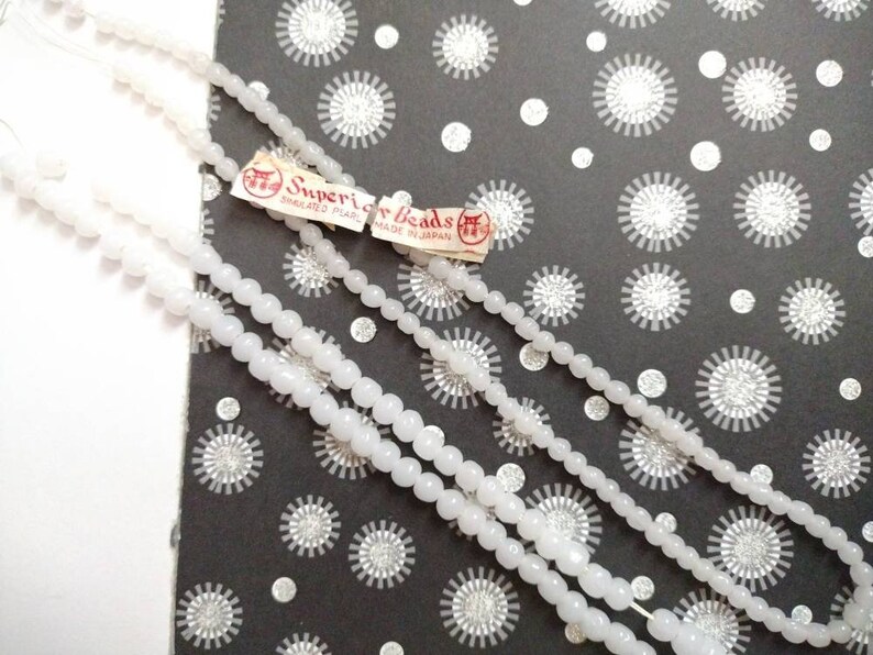 May include: Three strands of white pearl beads on a black and white patterned background. The beads are labeled "Superior Beads" and "Simulated Pearl Made in Japan."