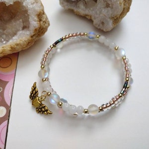 May include: A white and gold beaded bracelet with a gold angel wing charm. The bracelet is made with iridescent beads and small gold beads.