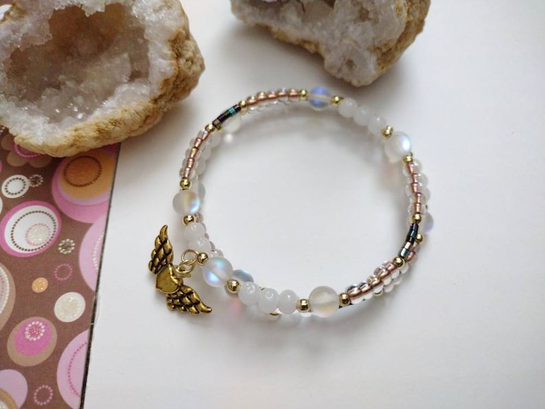 May include: A white and gold beaded bracelet with a gold angel wing charm. The bracelet is made with iridescent beads and small gold beads.
