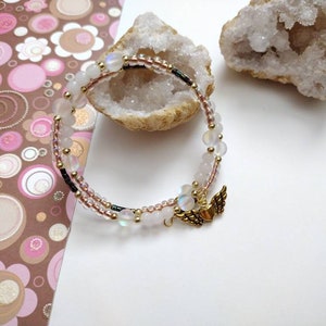 May include: A delicate white and gold bracelet with iridescent beads and a small gold charm with wings and a heart. The bracelet is on a white background.