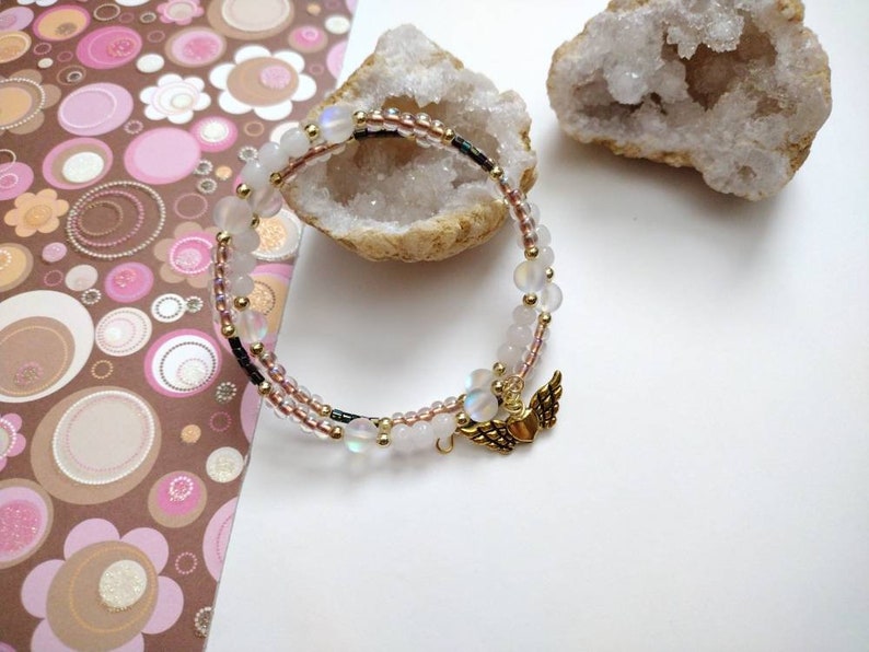 May include: A delicate white and gold bracelet with iridescent beads and a small gold charm with wings and a heart. The bracelet is on a white background.