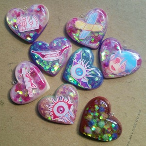 Puede incluir: Una colección de 10 dijes de resina en forma de corazón con varios diseños de purpurina y gráficos coloridos. Los diseños incluyen un cuchillo ensangrentado, una venda, un ojo espeluznante, una boca ensangrentada, un personaje de dibujos animados con una venda en el ojo y una palabra japonesa en rojo.