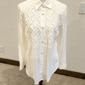 May include: Cream-colored button-down shirt with a woven design on the upper chest. The long-sleeved blouse has a classic collar and is displayed on a mannequin. The shirt is made of a soft, flowing fabric.