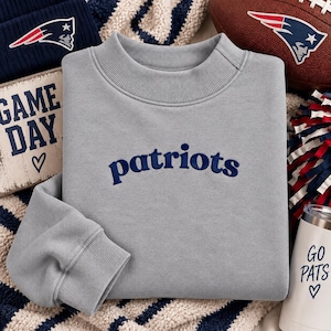 May include: A light gray crewneck sweatshirt featuring the word patriots embroidered in dark blue across the chest. The garment has a ribbed collar and ribbed cuffs on the long sleeves. The fabric has a smooth texture and a solid light gray color.
