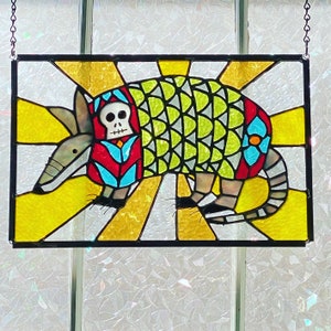 Könnte beinhalten: Ein hängendes Buntglasfenster mit einem farbenfrohen Gürteltier-Motiv. Das Gürteltier trägt eine rot-blau-weiß gemusterte Weste mit einem Totenkopf auf der Vorderseite. Der Hintergrund ist ein gelber Sonnenstrahl.