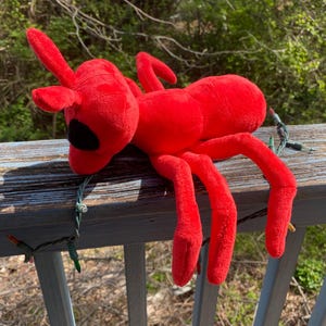 May include: A red plush ant toy with black eyes, lying on a wooden railing. The ant has six legs and is made of soft, fuzzy material.