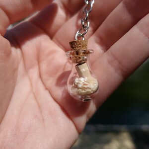 May include: A small glass bottle with a cork stopper, filled with sand and a seashell, hangs from a silver chain keychain.