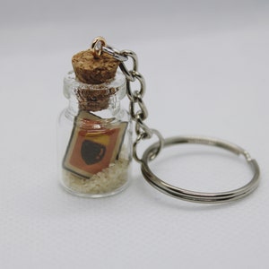 May include: A small glass bottle with a cork top and a silver chain attached. Inside the bottle is a small piece of paper with a red and yellow design and white sand.