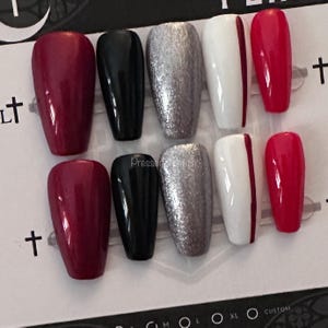 May include: A set of eight press-on nails in various colors and designs. The nails include solid burgundy, black, silver glitter, white with a burgundy stripe, and solid red. The nails are arranged on a white background.