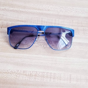 May include: Blue sunglasses with a textured frame and gradient lenses. The sunglasses have a rectangular shape with a double bridge and a dark tint. The brand name "JEAN-LOUIS SCHERRER PARIS" is visible on the frame.