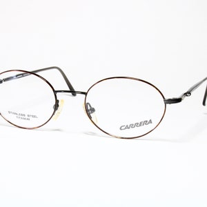 Puede incluir: Gafas ovaladas con montura marrón oscuro y varillas negras. Las lentes transparentes llevan el nombre de la marca "CARRERA" en la lente derecha. La montura es de acero inoxidable y titanio.