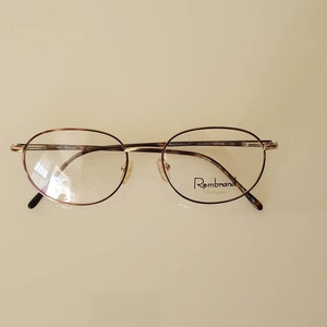 May include: Oval spectacles with a thin, rose gold metal frame. The lenses are clear, and the bridge features a nose pad. The brand name "Rembrandt" is printed on the right lens. The glasses are set against a light background.