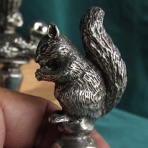May include: A silver-toned squirrel figurine with a bushy tail. The squirrel is holding a nut in its paws and is sitting on a base.