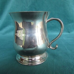 May include: A silver pewter tankard with a rounded body and a handle. The tankard has a smooth, polished surface.