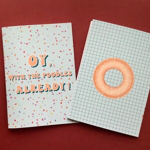 Puede incluir: Dos cuadernos con diseños diferentes. El primer cuaderno tiene un fondo blanco con confeti de colores y el texto "OY. WITH THE POODLES ALREADY!" El segundo cuaderno tiene un fondo blanco con un patrón de cuadrícula y una rosquilla de color melocotón.