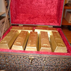 May include: A wooden treasure chest with a red velvet lining is open and filled with gold bars. The chest has intricate carvings and a metal latch.