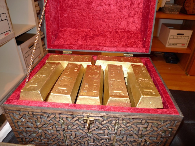 May include: A wooden treasure chest with a red velvet lining is open and filled with gold bars. The chest has intricate carvings and a metal latch.