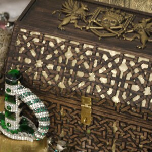 May include: A wooden treasure chest with intricate carvings and a brass lock. The chest is decorated with a skull and crossbones and two griffins. The chest is surrounded by gold coins and a jeweled dagger.