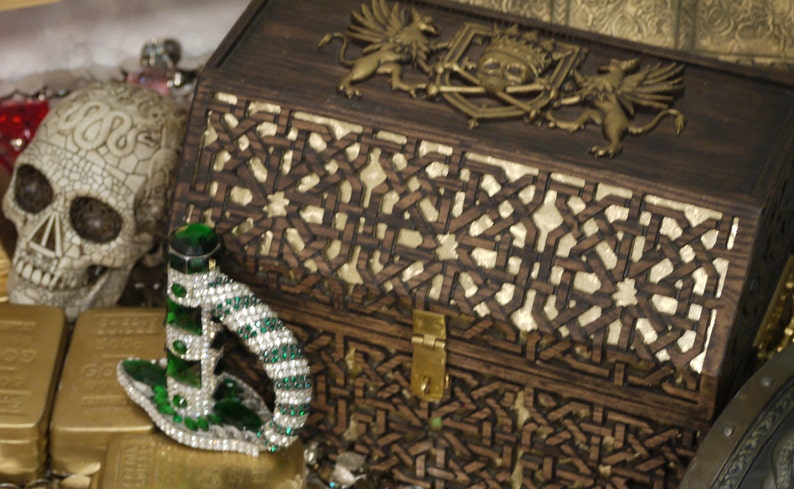 May include: A wooden treasure chest with intricate carvings and a brass lock. The chest is decorated with a skull and crossbones and two griffins. The chest is surrounded by gold coins and a jeweled dagger.