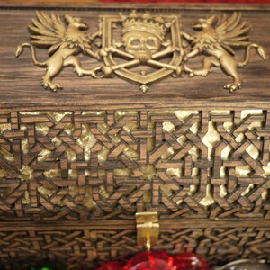 May include: A wooden box with a gold-colored skull and crossbones design on the lid. The box is decorated with intricate carvings and has a gold-colored latch.