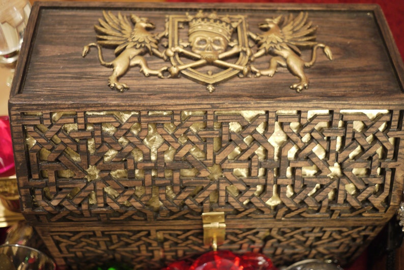 May include: A wooden box with a gold-colored skull and crossbones design on the lid. The box is decorated with intricate carvings and has a gold-colored latch.