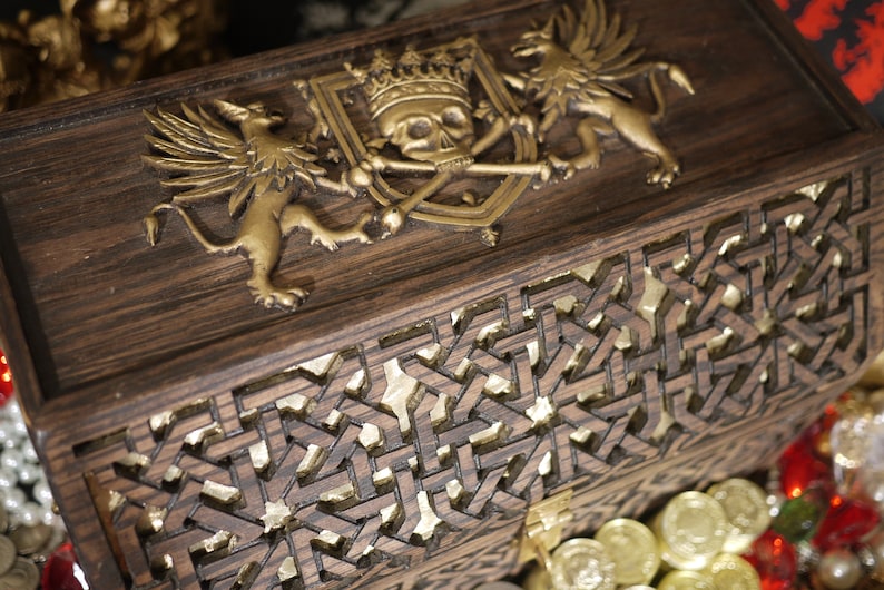 May include: A wooden treasure chest with a gold-colored skull and crossbones design on the lid. The chest is decorated with intricate carvings and has a gold-colored latch.