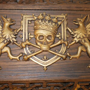 May include: A gold-colored wooden plaque with a carved design of a skull wearing a crown, flanked by two winged griffins.