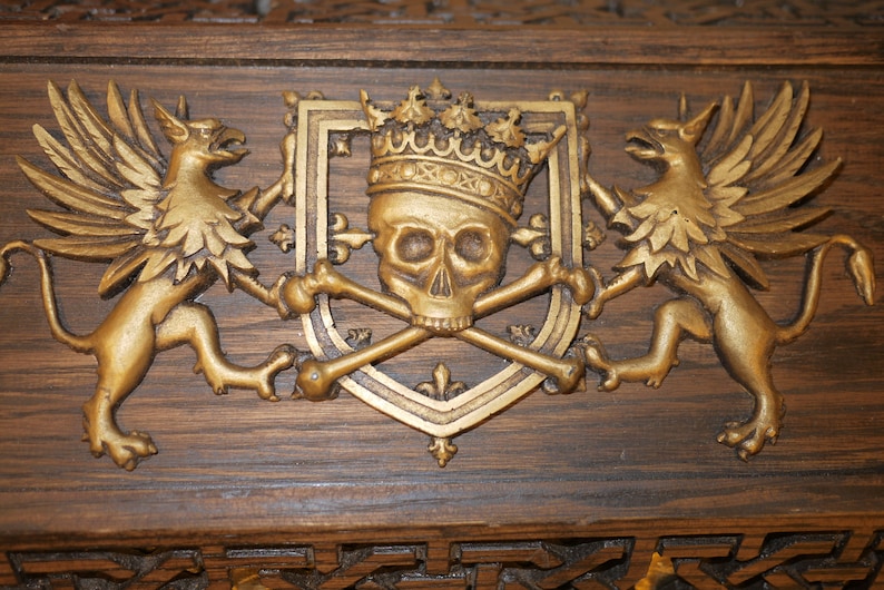 May include: A gold-colored wooden plaque with a carved design of a skull wearing a crown, flanked by two winged griffins.