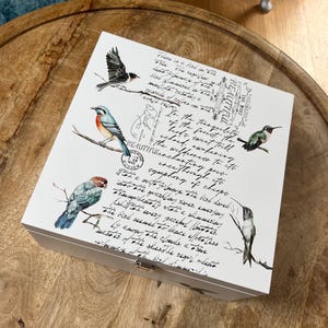 May include: White wooden box with a hinged lid, decorated with a vintage-inspired design featuring birds and a handwritten quote. The quote reads: "There is a bird in the tree...the result of the first sweet trill echoed, nantering to the wilderness to its enchanting pres...a symphony of chirps."