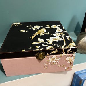 May include: A decorative wooden box. The black lid features a gold bird and floral design. The box has a pink base with matching floral accents and a brass clasp. The box is a square shape.