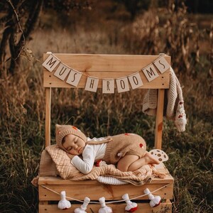 Puede incluir: Un bebé con un mono marrón con temática de champiñones duerme en una caja de madera con un letrero que dice "Champiñones". La caja está decorada con una cadena de adornos blancos con forma de champiñones.