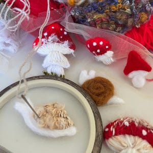 May include: An assortment of handmade felted mushrooms with red caps and white spots, along with a snail and embroidery hoop. The mushrooms vary in size and shape, and are made with white and red yarn.
