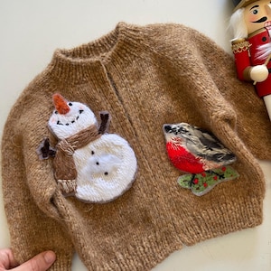 Puede incluir: Cárdigan de punto marrón con apliques de muñeco de nieve y pájaro. El muñeco de nieve tiene una nariz de zanahoria y una bufanda marrón. El pájaro es rojo, gris y negro. Un cascanueces de madera está en el fondo.