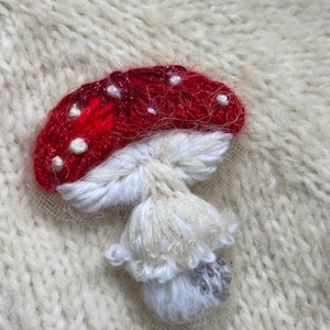 Puede incluir: Un hongo bordado de color rojo y blanco con puntos blancos en el sombrero, cosido sobre una tela de punto blanca.