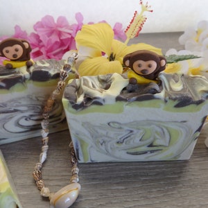 May include: Two bars of handmade soap with a swirl pattern of green, white, and brown. The soap is decorated with small monkey figurines and a shell necklace.