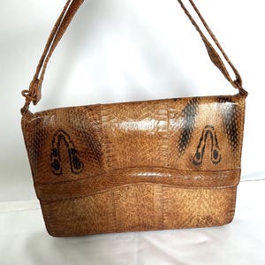 May include: A rectangular shoulder bag featuring a textured, light brown snakeskin pattern with darker brown, U-shaped markings on the front flap. The bag includes a matching thin shoulder strap attached to the sides with small metal hardware.