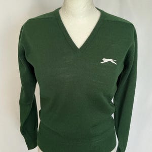 May include: A dark green V-neck jumper with long sleeves. A white leaping animal logo is on the left chest. The jumper is displayed on a white mannequin.