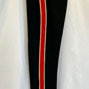 Op de afbeelding: Zwarte broek met een rode en crèmekleurige streep aan de zijkant. De broek heeft een ritssluiting en een zak. De broek hangt aan een hanger tegen een witte achtergrond.