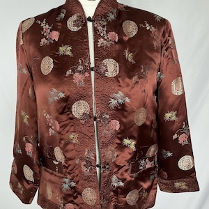 May include: A dark brown satin jacket featuring a mandarin collar and long sleeves. The garment is decorated with an all-over pattern of circular medallions, floral motifs, and bamboo stalks. It closes at the front with black fabric toggle buttons.