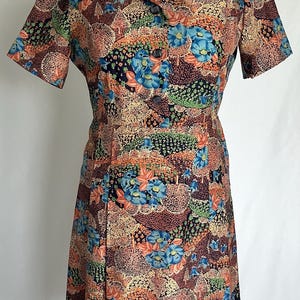 May include: A vintage dress with a floral pattern. The dress has short sleeves, a collared neckline, and a button-down front. The fabric displays a pattern of blue, orange, and green flowers on a brown background. The dress is displayed on a mannequin.