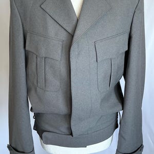 May include: A grey military-style jacket with a tailored fit. The jacket has two front pockets, shoulder epaulettes, and buttoned cuffs. The jacket is displayed on a white mannequin against a white backdrop.